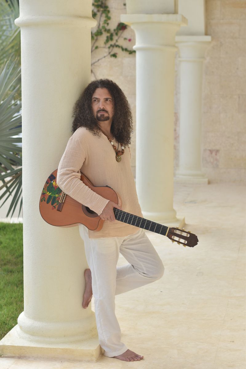 Photo of a man in white, holding a guitar and leaning against a white pillar